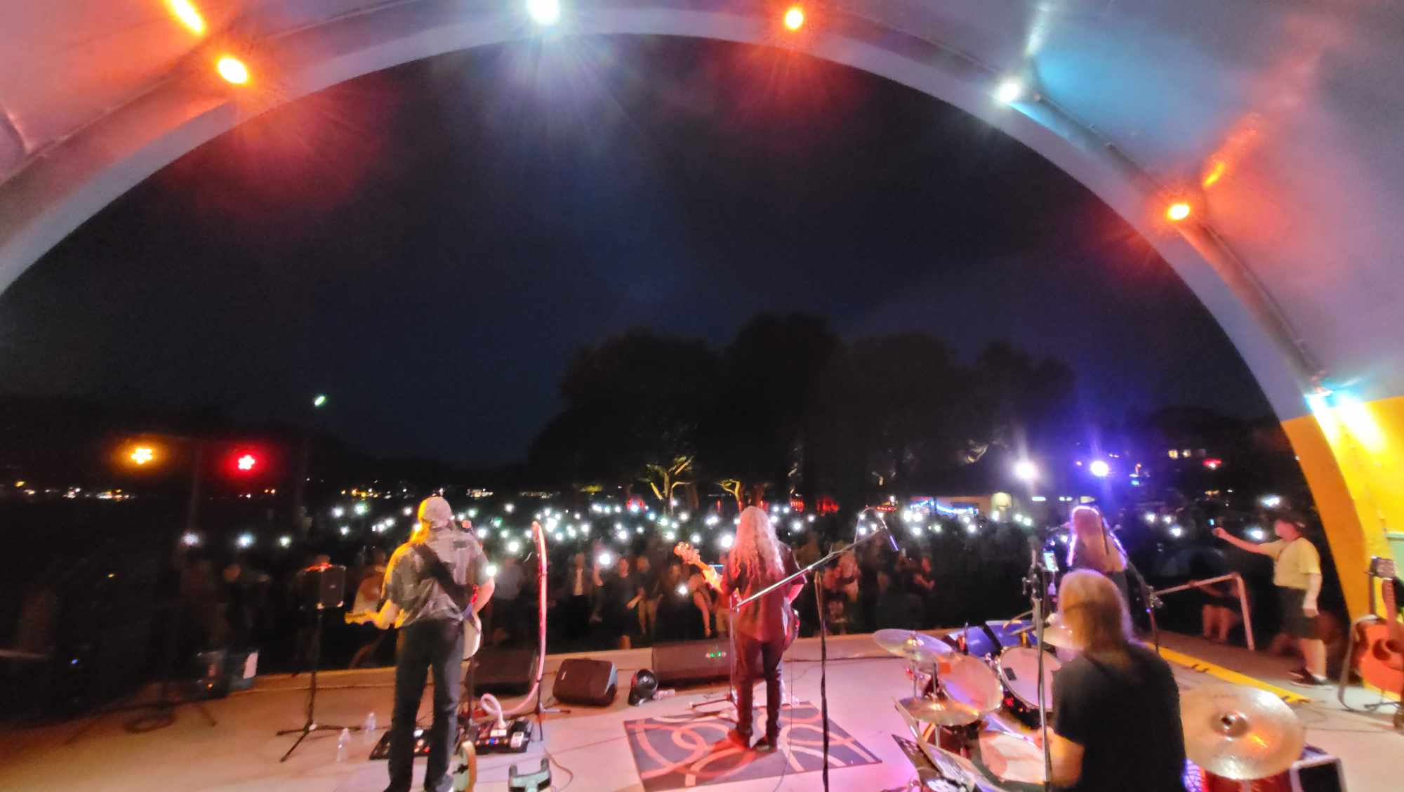 The Eagle Eyes performing live on an outdoor bandshell stage at night, facing a crowd illuminated by cell phone lights